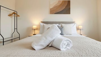 Un lit bien fait, avec du linge de lit blanc et des serviettes pliées, deux lampes de chevet, un cintre en bois et un tableau accroché au mur.