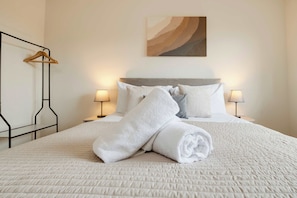 A neatly made bed with white linens and rolled towels, two bedside lamps, a wooden hanger, and a painting on the wall.