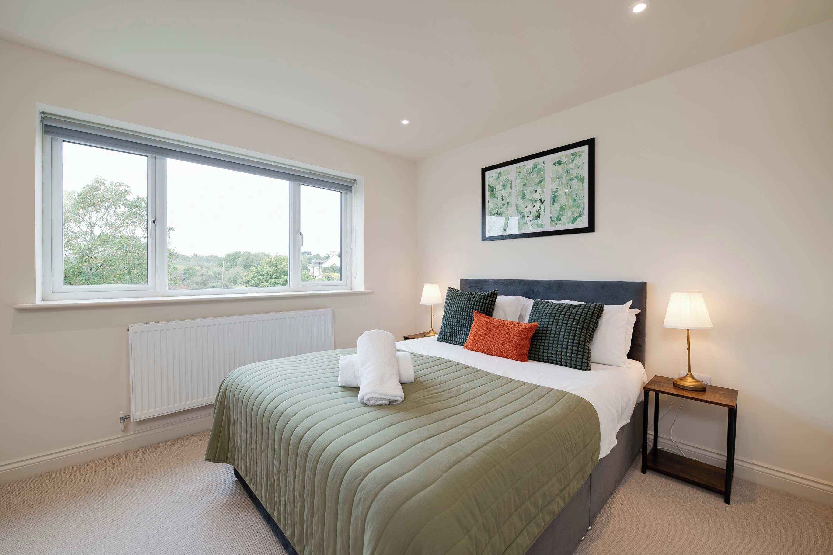 A hotel room with a large bed, a window with blinds, a radiator, a bedside table with a lamp, and a framed picture on the wall.