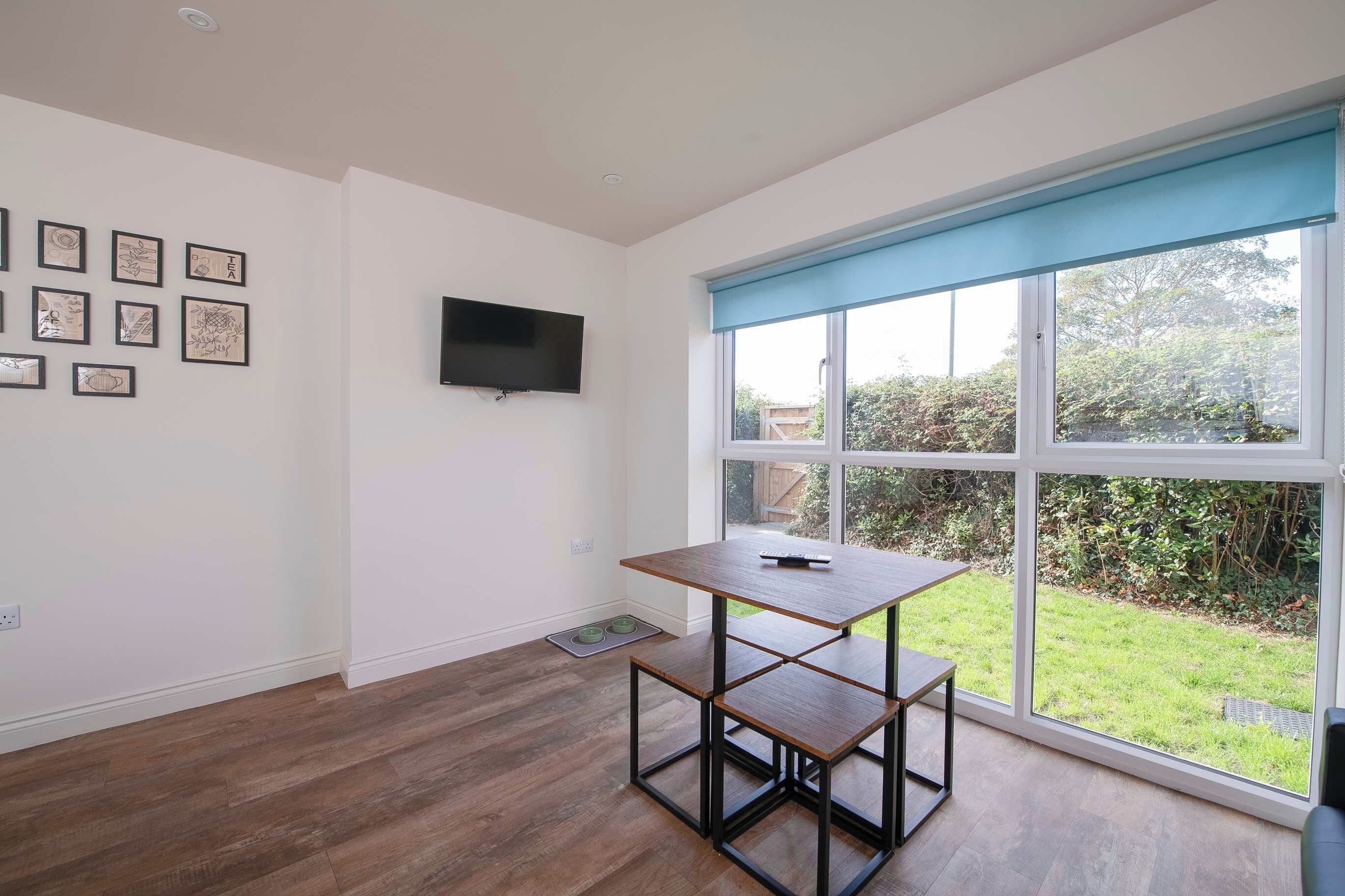 A room with a large window, a flat-screen TV mounted on the wall, and a wooden table with a remote control on it.