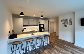 Una cocina moderna con una isla central, taburetes de bar, iluminación colgante y un refrigerador.