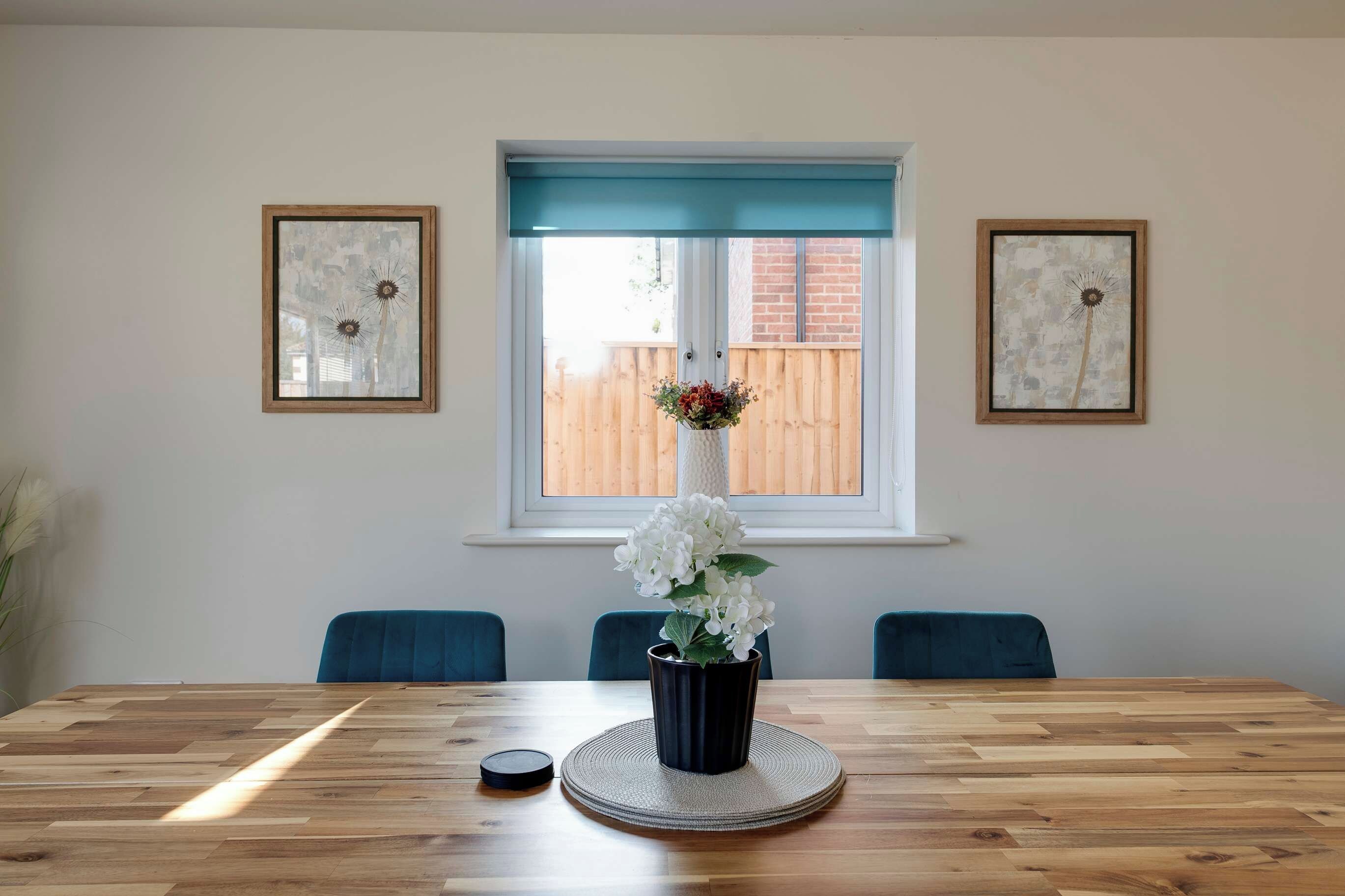 A dining area with a wooden table, chairs, a vase with flowers, and two framed pictures on the wall.
