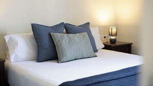 Una cama bien hecha con sábanas blancas y dos almohadas, una lisa y otra a rayas, en una habitación con una mesita de noche y una lámpara.