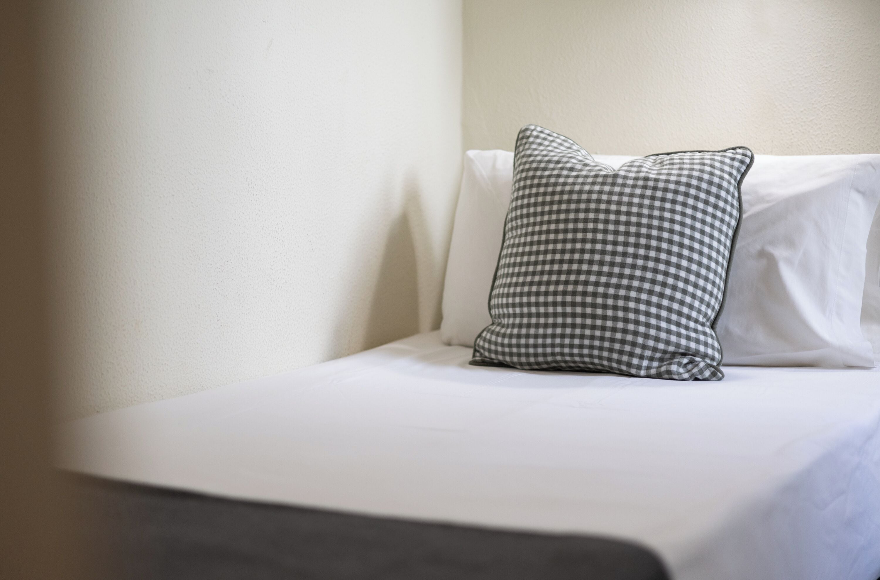 Un lit avec un oreiller à carreaux et un oreiller blanc uni, le tout placé contre un mur uni.