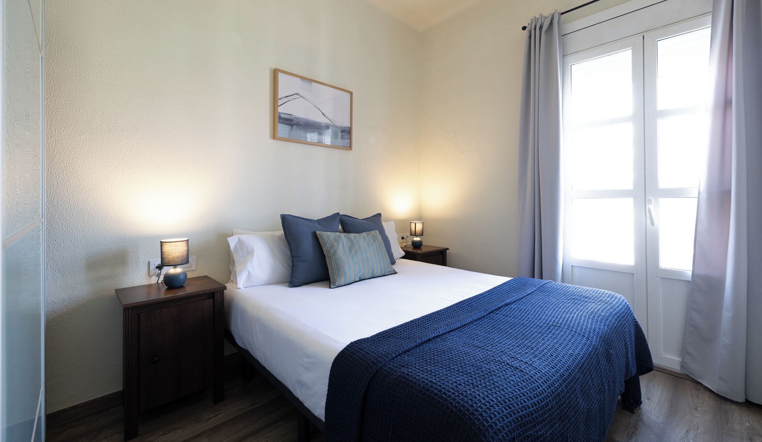 Habitació d'hotel amb un llit, dues taules de nit, una tauleta de nit, una làmpada, una finestra amb cortines i un quadre penjat a la paret.