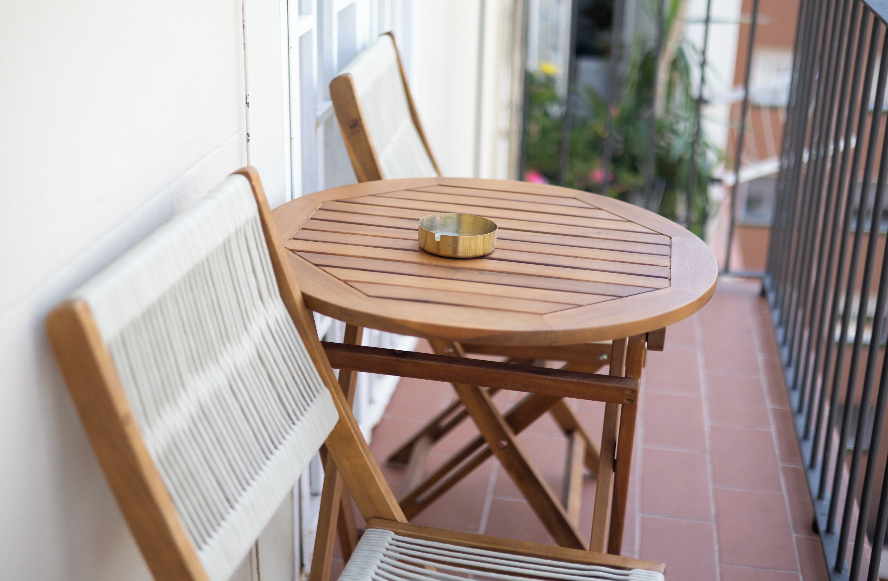 A round wooden table with a metal ashtray on a balcony with a metal railing.