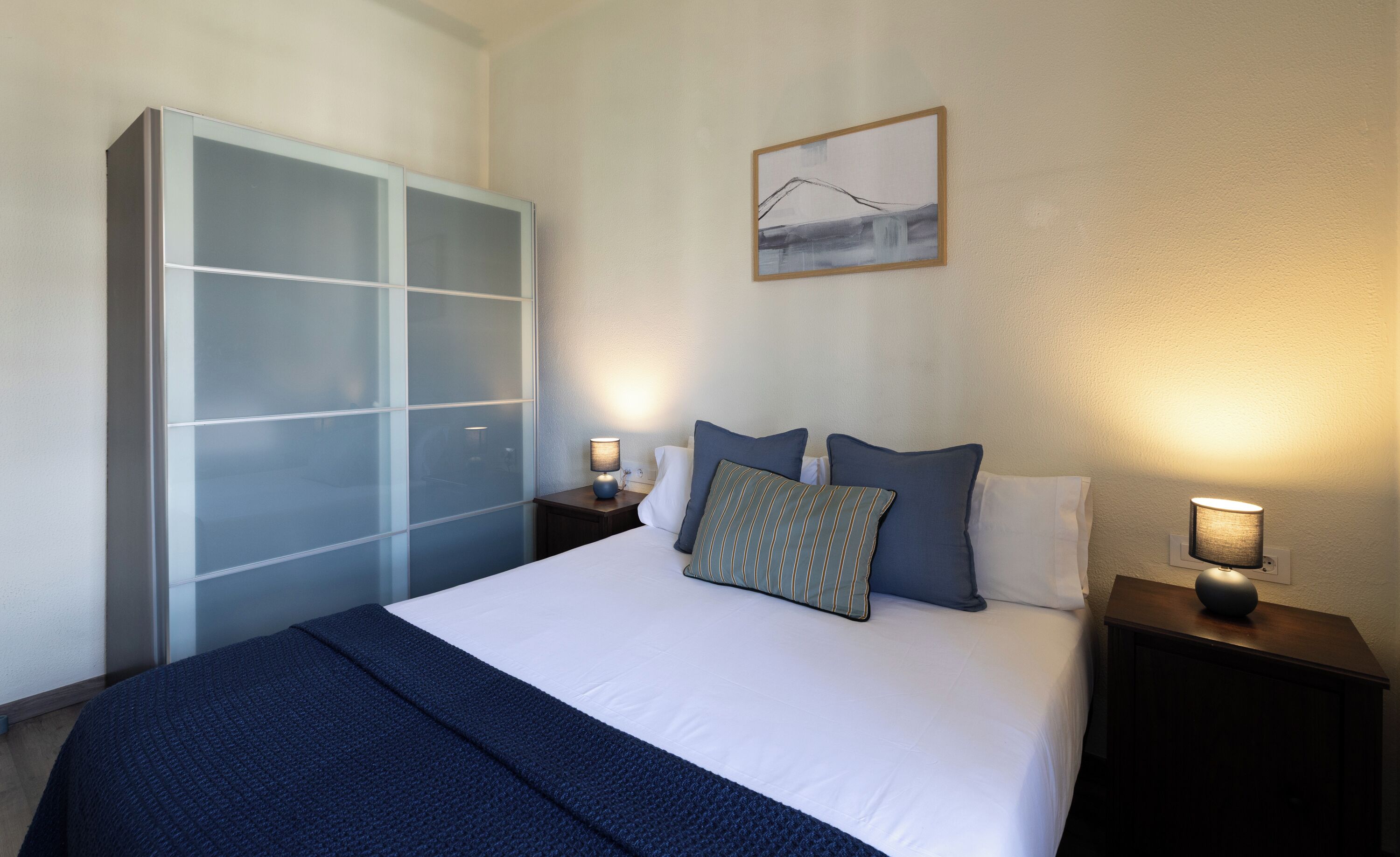 A hotel room with a bed, a nightstand with a lamp, a wardrobe, and a painting on the wall.