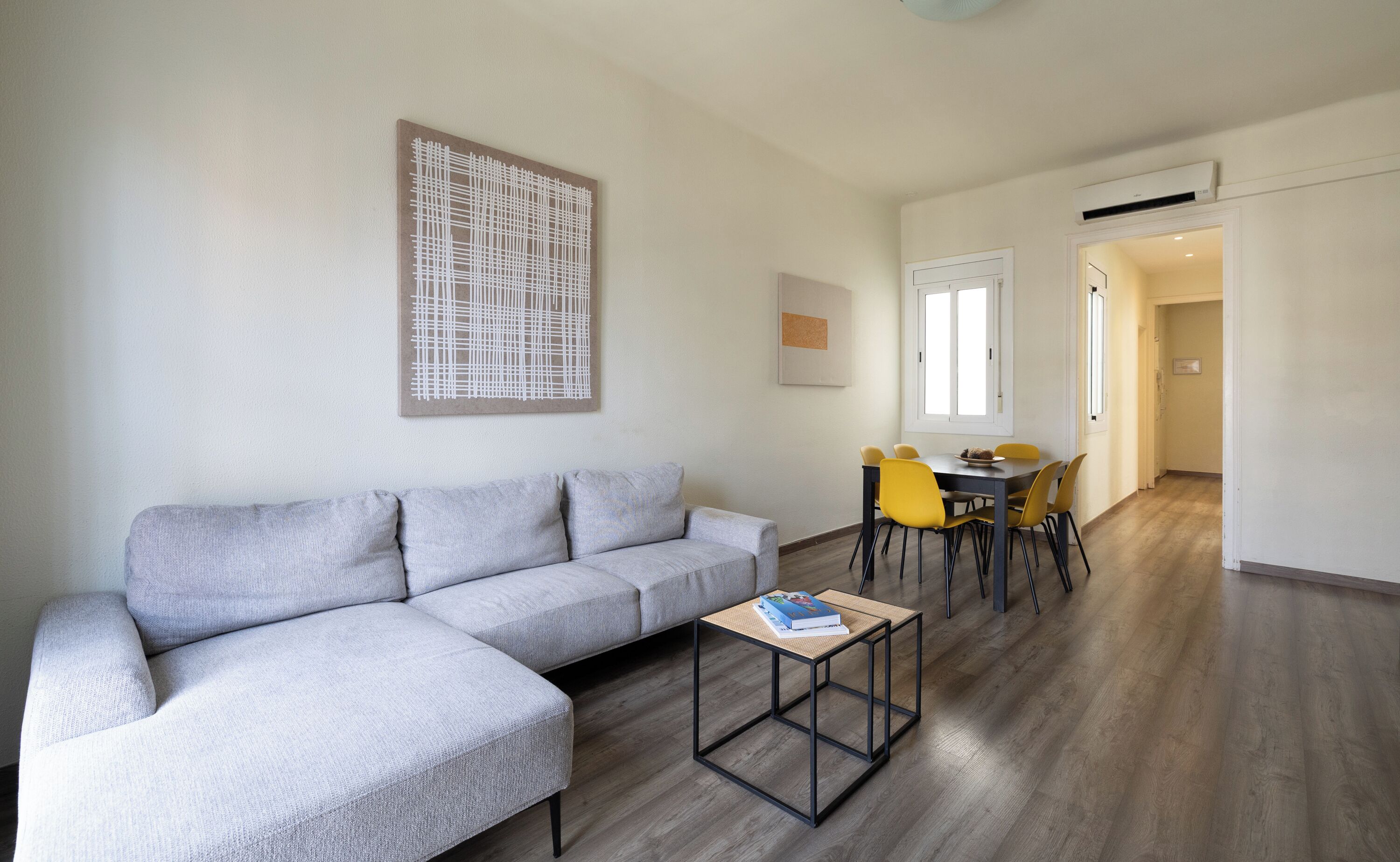 Un salon moderne avec un canapé d’angle gris, une petite table basse et un coin repas avec des chaises jaunes.