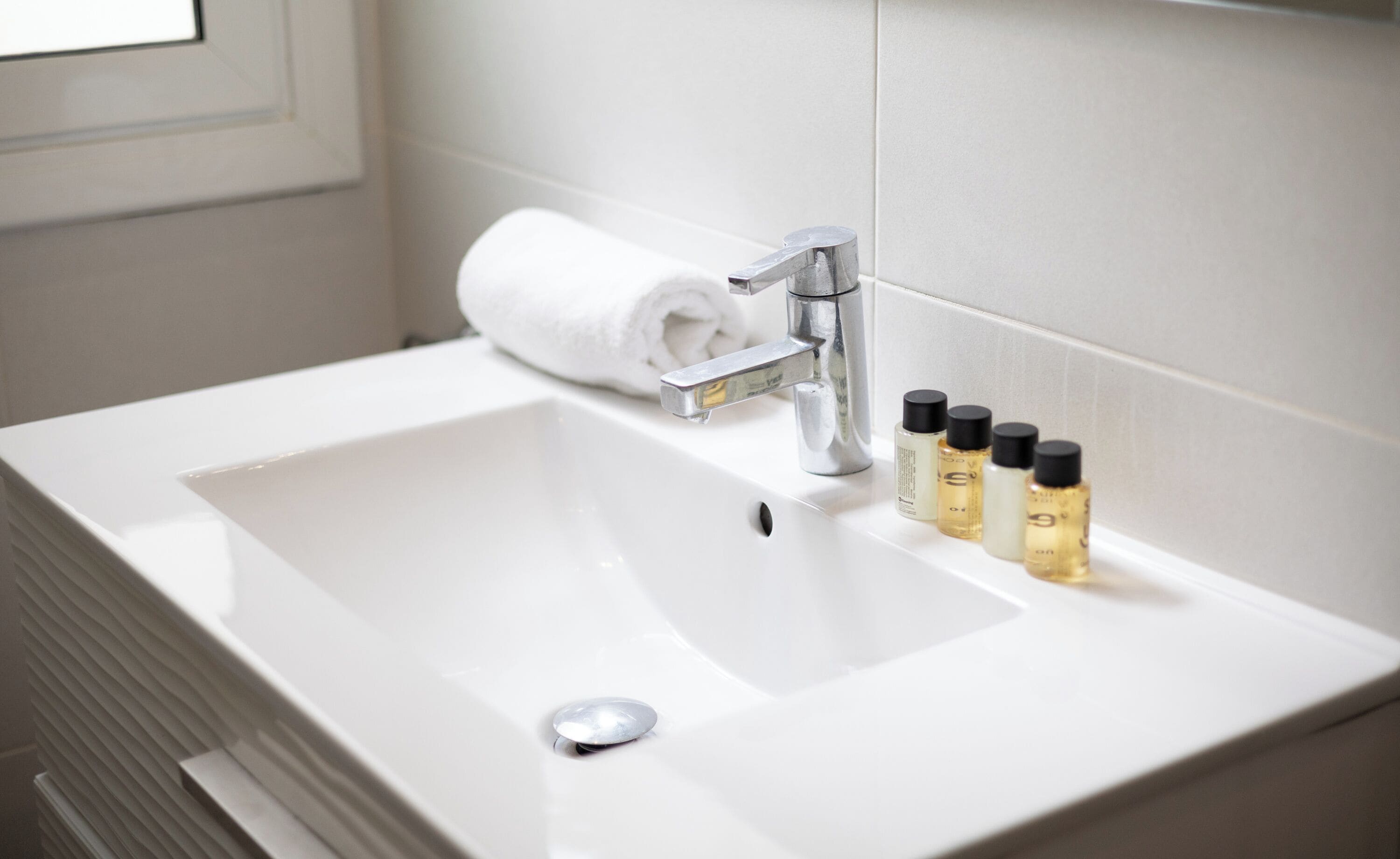 A white bathroom sink with a modern faucet, a rolled white towel, and four bottles of toiletries.