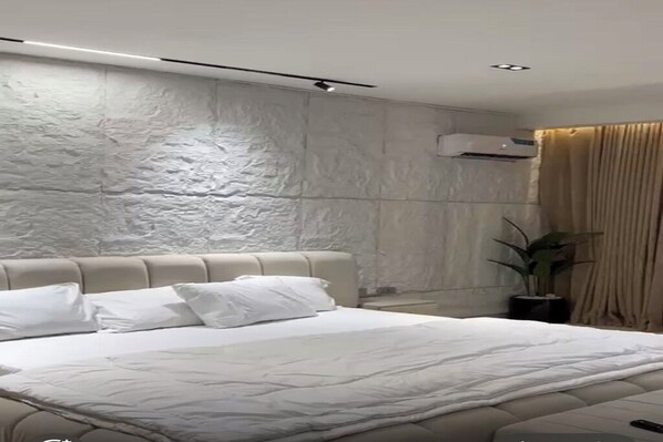 A modern bedroom with a large bed, a headboard, an air conditioning unit, a potted plant, and a window with curtains.