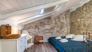 Ein kleiner, gemütlicher Raum mit schräge Decke, einer Steinwand, einem Bett mit blauer Bettwäsche, einer hölzernen Kommode und einem Stuhl.