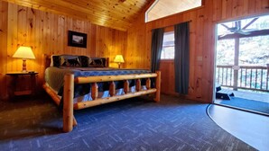 Ein Schlafzimmer einer Holzhütte mit einem Bett, zwei Nachttischlampen, einem Fenster mit Vorhängen und Blick ins Freie.