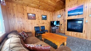 Ein Wohnzimmer mit Holzwänden, einem braunen Ledersofa, zwei blauen Sesseln, einem Holztisch und einem Kamin. Ein Flachbildfernseher ist ebenfalls vorhanden.