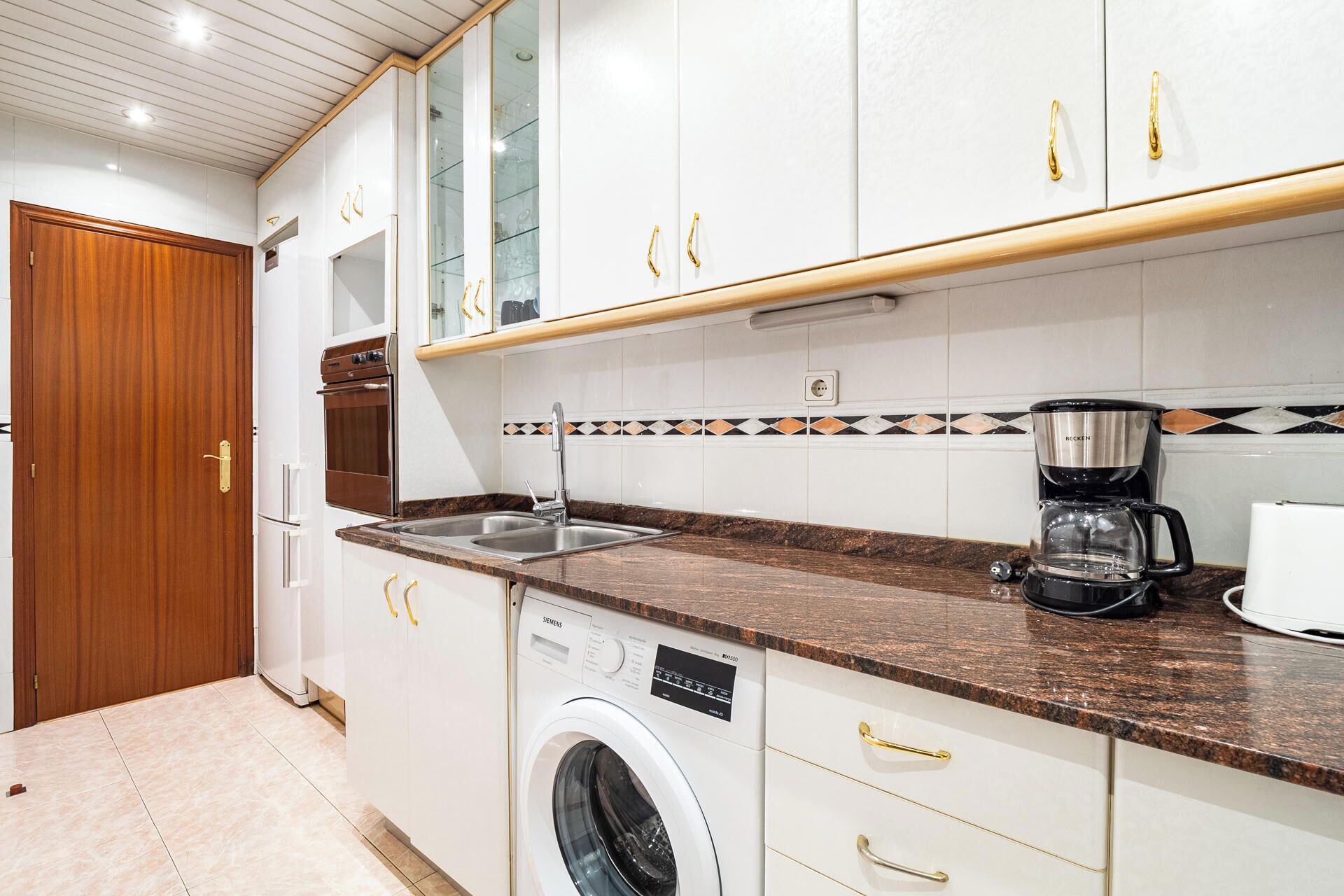 A compact kitchen with white cabinets, a built-in washing machine, a coffee maker, and a wooden door.