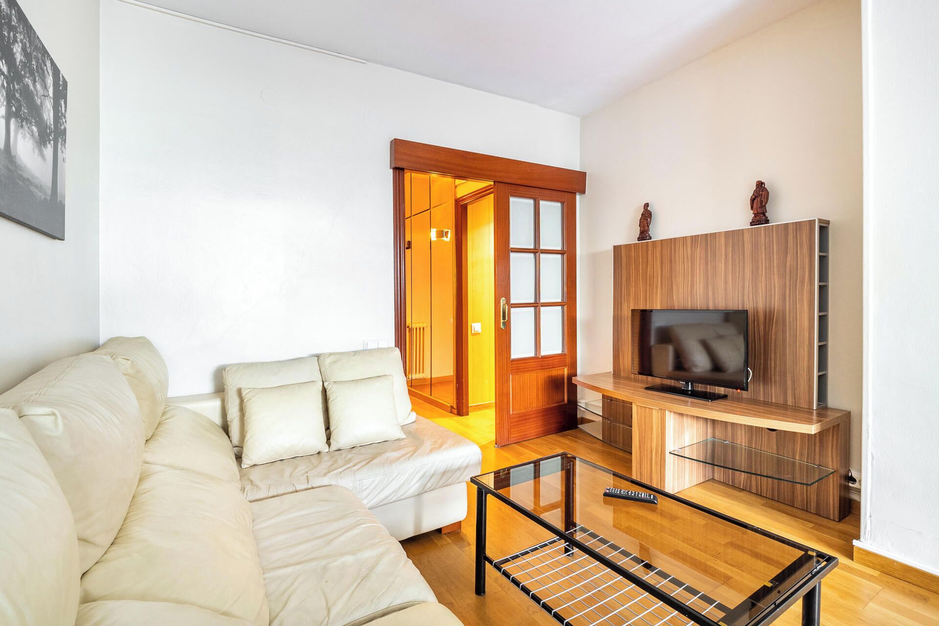 A living room with a sofa, a glass coffee table, a TV on a wooden stand, and a wooden door leading to another room.