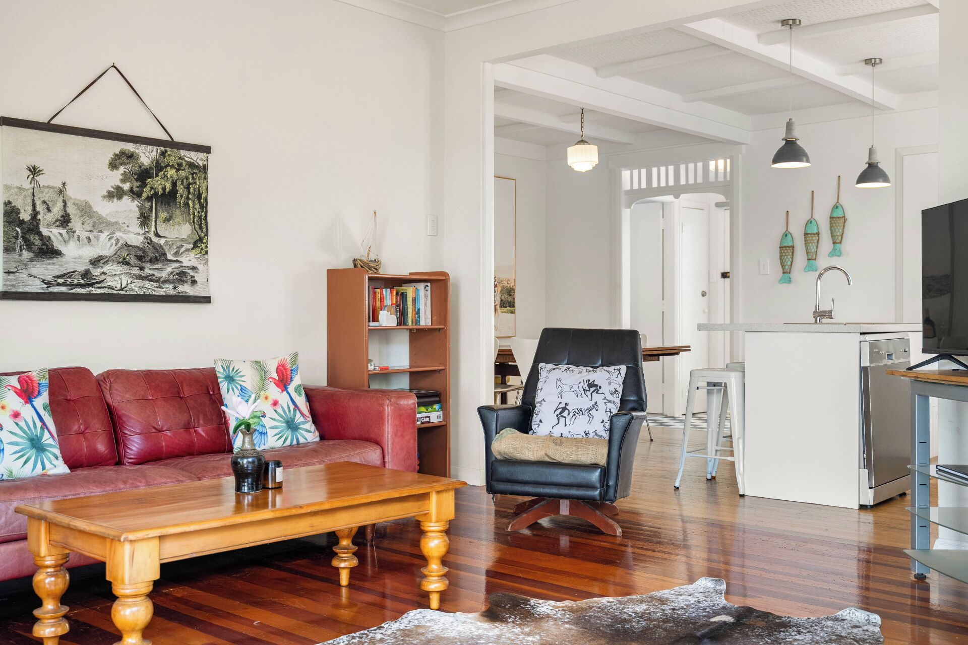 A living room with a red sofa, a wooden coffee table, a black armchair, a bookshelf, and a kitchen area with a refrigerator and a sink.