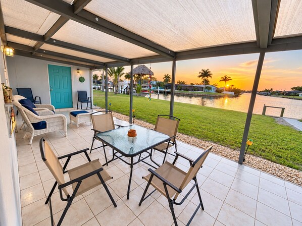 A covered outdoor seating area with a table and chairs, overlooking a body of water and a sunset.