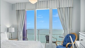 A hotel room with a bed, a chair, a dresser, and a view of the ocean through a sliding glass door. - Ocean Front Oasis; Stunning 2BR with Private Double Balcony at The Wyndahm Ocean Walk Resort #2221 (Daytona Beach)