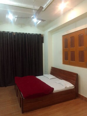 A room with a wooden bed, a red blanket, a ceiling fan, and a window with wooden shutters. - Maa Annapurna grand guests house (Varanasi)