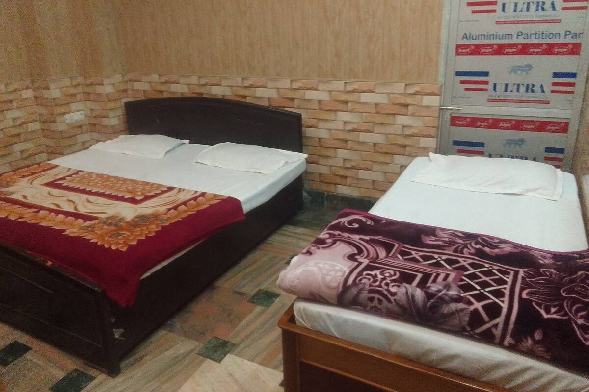 Una habitación con dos camas, una con colcha roja y beige y otra con colcha púrpura y blanca. Hay un cartel en la pared que anuncia 'ULTRA Aluminium Partition Pan'.