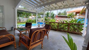 Un espace extérieur aménagé avec des meubles en osier, abrité et offrant une vue sur une piscine et un jardin luxuriant.