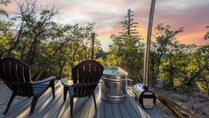A wooden deck with two Adirondack chairs and a metal table, surrounded by trees and a sunset in the background. - Bryce and Zion middle point with Cowboy Hot tub (Alton)