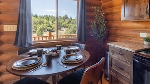Ein Holzblockhaus-Interieur mit einem für vier Personen gedecktem Esstisch, einem Blumentopf und Blick auf den Wald durch große Fenster.