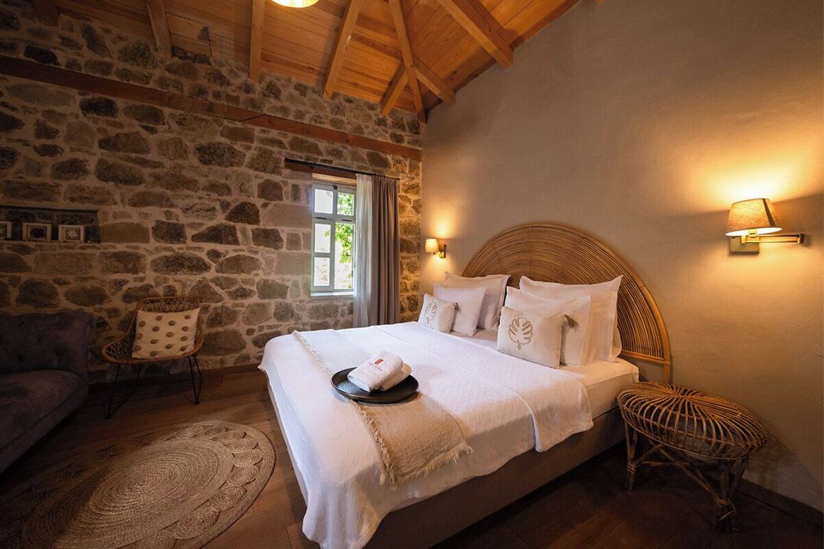 A bedroom with a stone wall, wooden ceiling beams, a bed with white linens, a wicker chair, and a small round table.