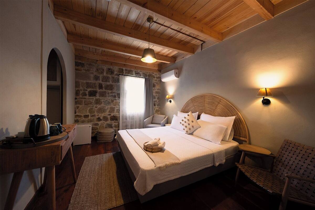 A cozy bedroom with a stone wall, a wooden ceiling, a bed with white linens, a bedside table with a kettle, and a chair.