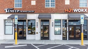 En byggnad med flera butikslokaler, inklusive ”TuneUp The Manly Salon” och ”HotWorx”. Det finns skyltar som visar var det finns reserverade parkeringsplatser och information om ett VIP-program.