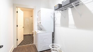 Ett tvättrum med en tvättmaskin och en torktumlare placerade ovanpå varandra, en vit tvättkorg och en metallhylla med korgar.