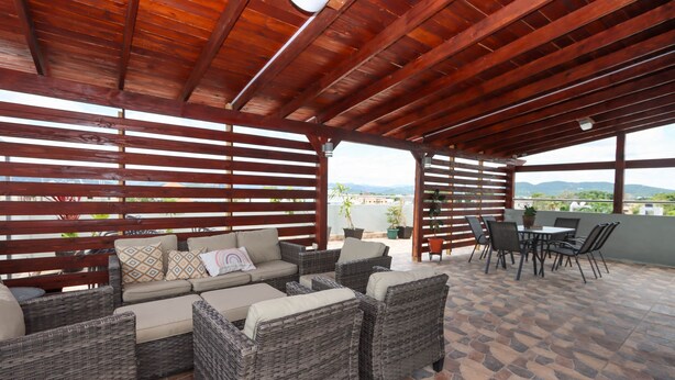 Un espacio de estar al aire libre con techo, con mobiliario de mimbre y una mesa, con vistas a un paisaje pintoresco.