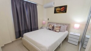 A bedroom with a bed, two bedside tables, a nightstand, a window with curtains, an air conditioner, and a wall-mounted painting. - Unique 2BR/2BA Apt – Stylish, Modern & High-End (San Cristobal)