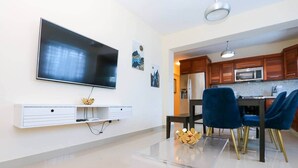 A modern living room with a flat-screen TV, a white entertainment unit, a dining area with blue chairs, and a kitchen with wooden cabinets.