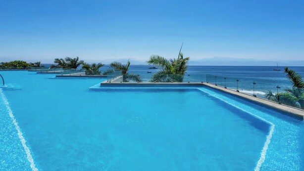 Una piscina grande y cristalina con vistas al océano y montañas lejanas.
