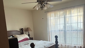 Un dormitorio con cama de madera, ventilador de techo, ventana con cortinas traslúcidas y lámpara en una mesita de noche.