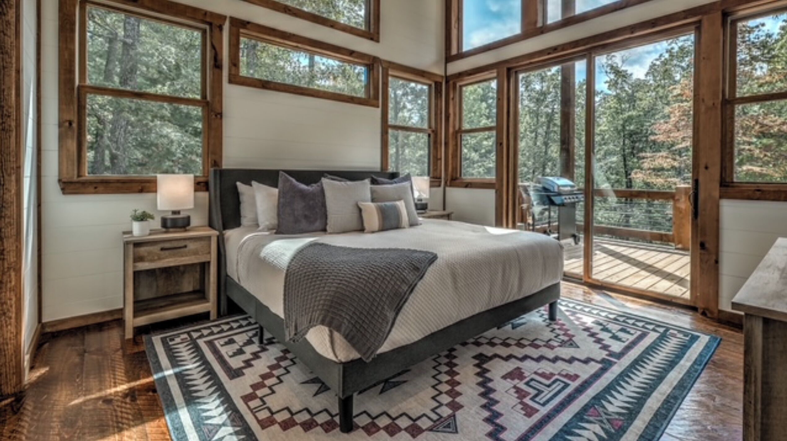 A bedroom with a large bed, wooden headboard, and a view of the outdoors through large windows.