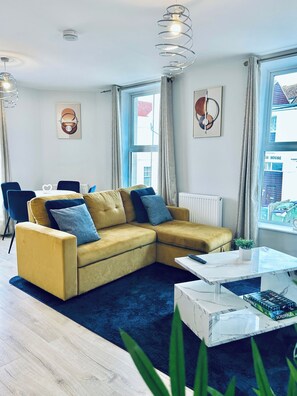 A modern living room with a yellow sofa, a white coffee table, and a blue rug. There are two pendant lights, a radiator, and a window with curtains.