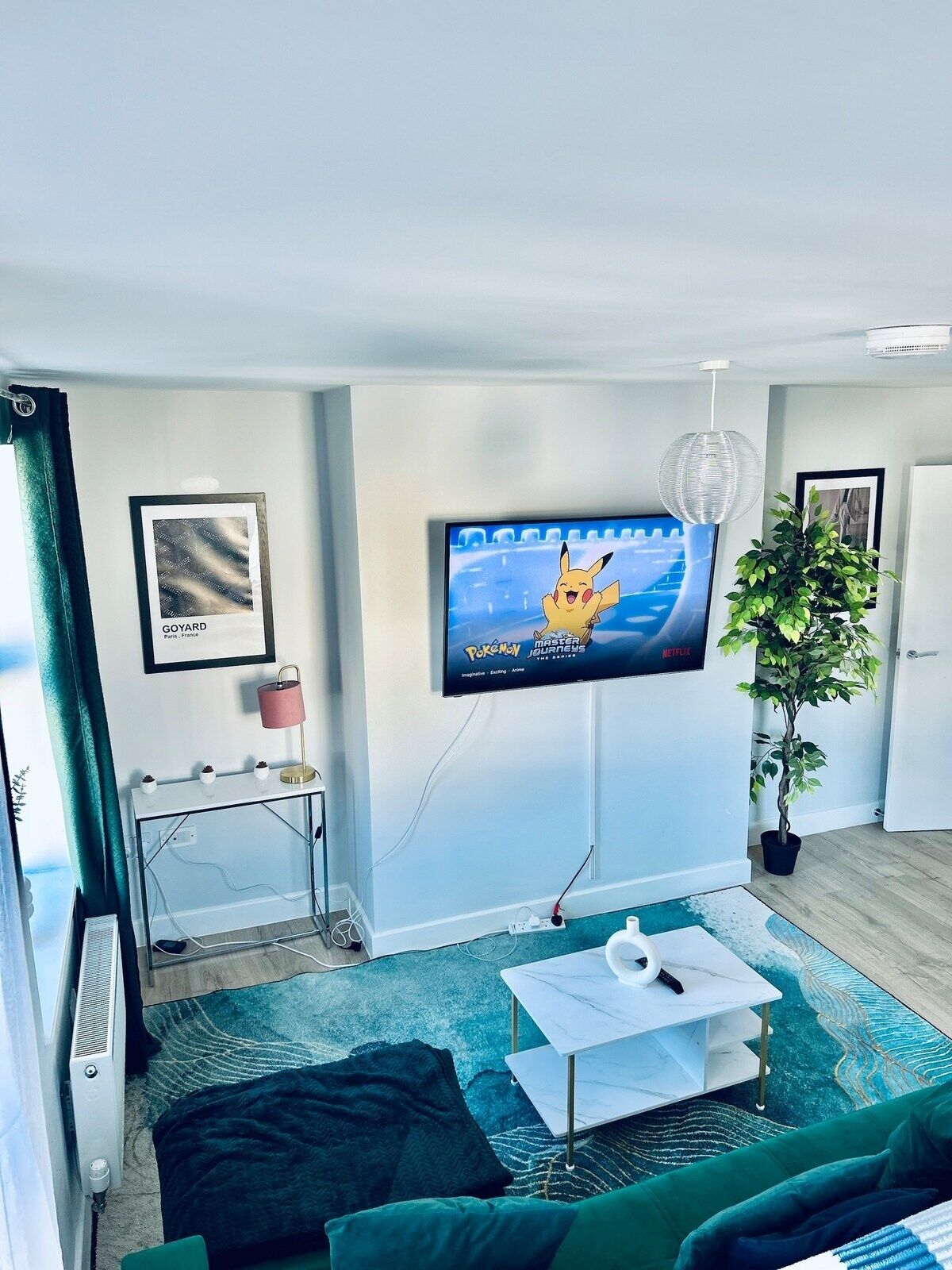 A modern living room with a flat-screen TV displaying 'Pokémon Master Journeys', a glass coffee table, a green sofa, a potted plant, and a framed map of Paris.