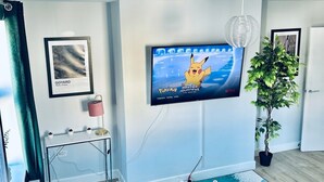 Una sala de estar moderna con un televisor de pantalla plana mostrando 'Pokémon Master Journeys', una mesa de centro de vidrio, un sofá verde, una planta en maceta y un mapa enmarcado de París.