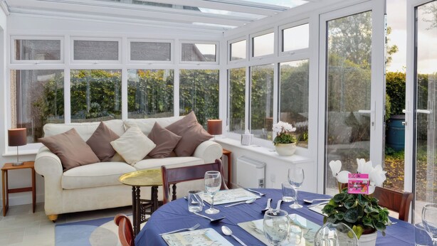 A conservatory with a dining table set for two, a sofa, and a view of greenery outside.