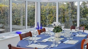 A dining table set for six with a blue tablecloth, wooden chairs, and a view of greenery through large windows.