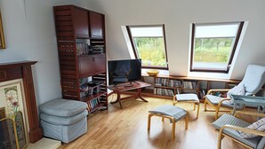 A living room with a wooden bookshelf, a television, and two windows with a view of greenery.
