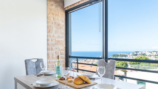 A dining table set with plates, cutlery, glasses, and a croissant, with a bottle of olive oil and nuts, overlooking a cityscape through a large window. - Pena View Apartment by Atlantic Holiday (Funchal)