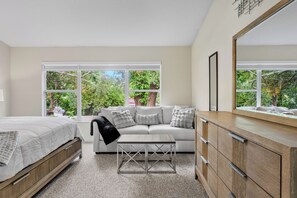 Un dormitorio con cama, sofá, una mesita de centro y cómoda. Amplias ventanas ofrecen vista a árboles.