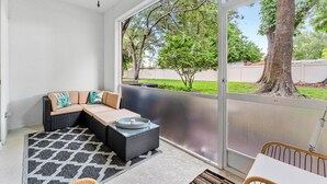 Un patio soleado con zona de estar con cojines, una pequeña mesa y vistas a un jardín arbolado.