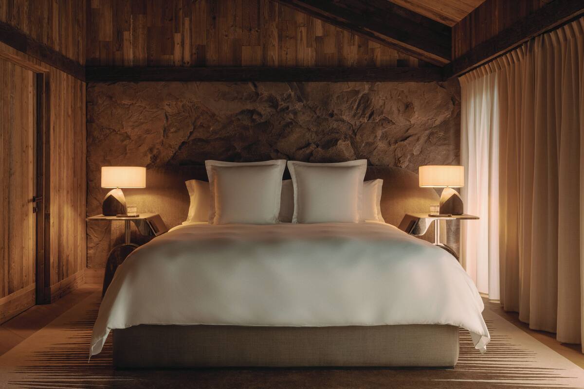 A large bed with a wooden headboard, two bedside tables with lamps, and a stone wall behind the bed.