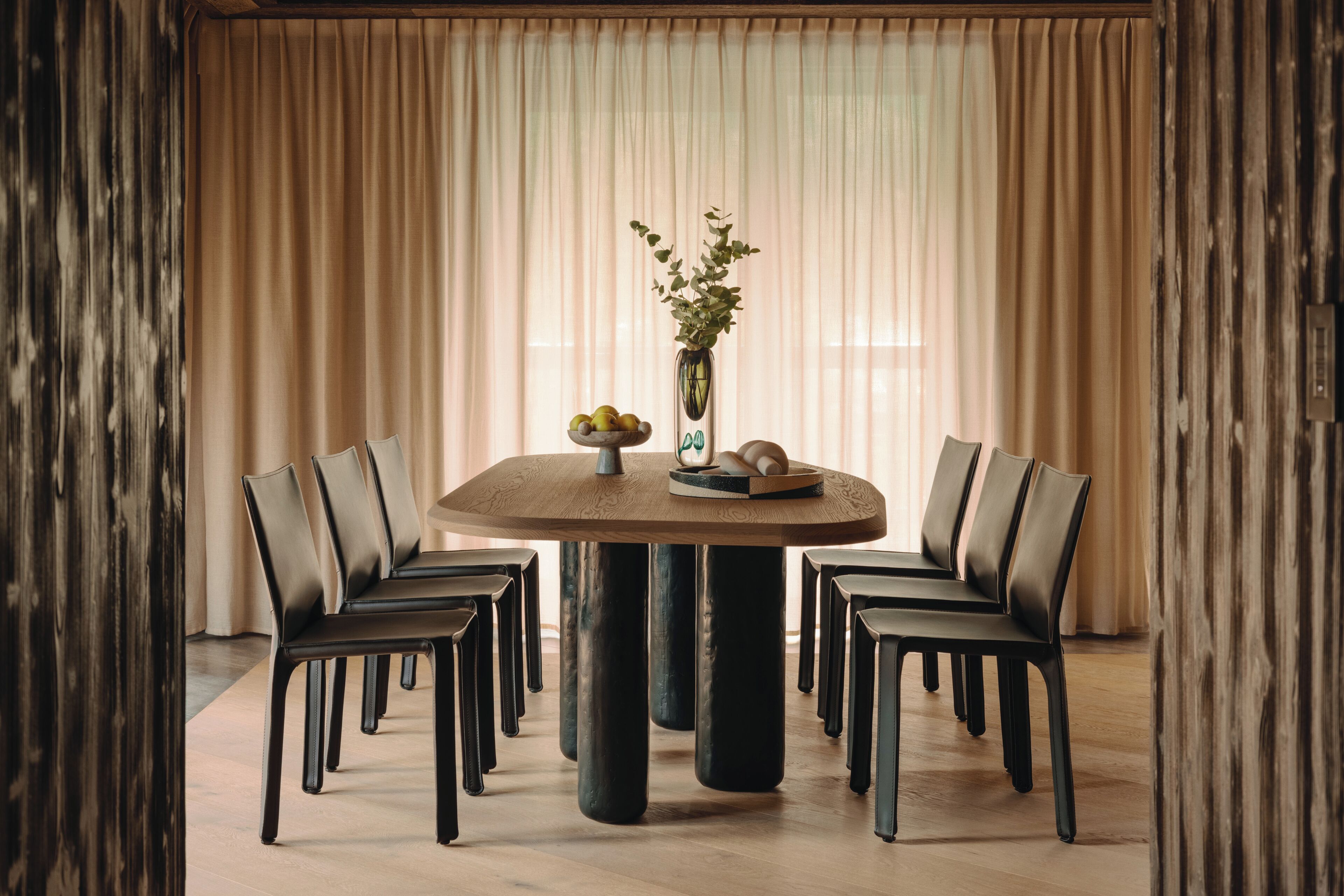A round dining table with six chairs, a vase with flowers, and a bowl of fruit on the table.