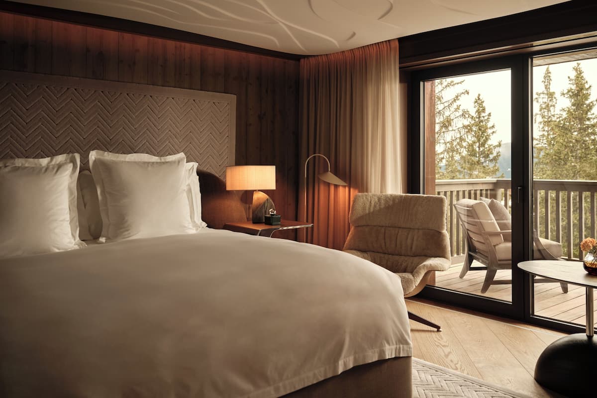 A hotel room with a large bed, a chair, a table, and a balcony with a view of trees.