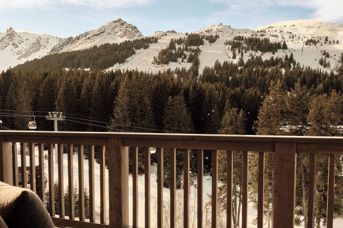 A wooden deck with a bench, overlooking a snowy mountain landscape with trees and a ski lift.